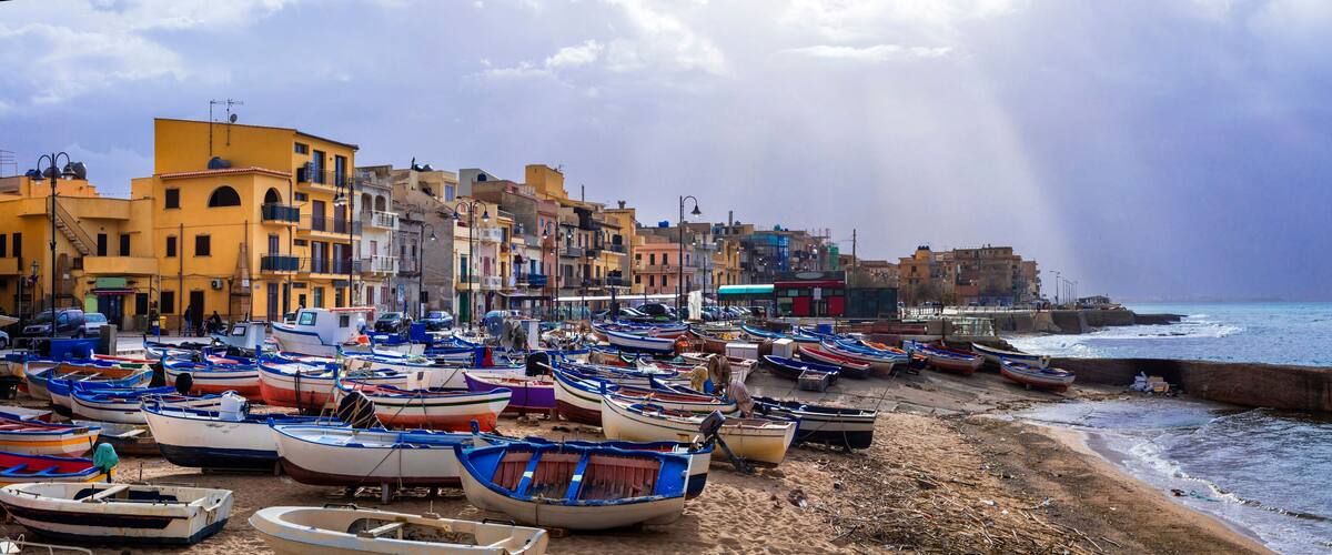 A coastal town with colorful boats docked along the shore, buildings in the background, and a stormy sky.