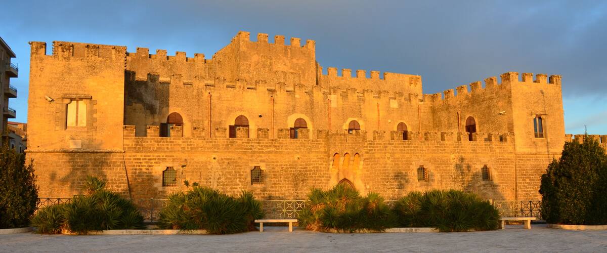 A historic stone castle with arched windows and battlements, surrounded by a paved courtyard and greenery.
