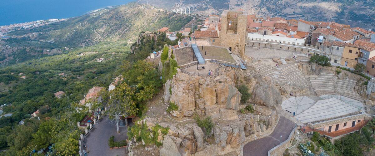 A coastal town with a historic amphitheater, terraced houses, and a bridge in the background.