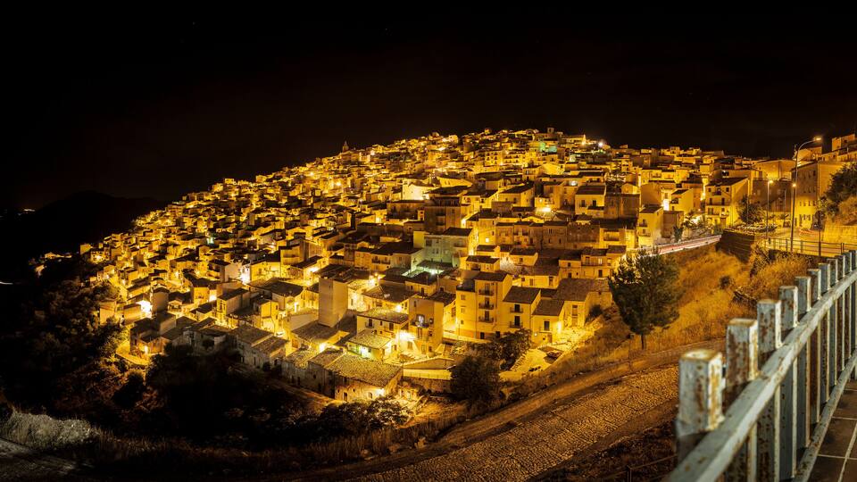 A night view of a densely packed hillside town with illuminated buildings and a railway track.