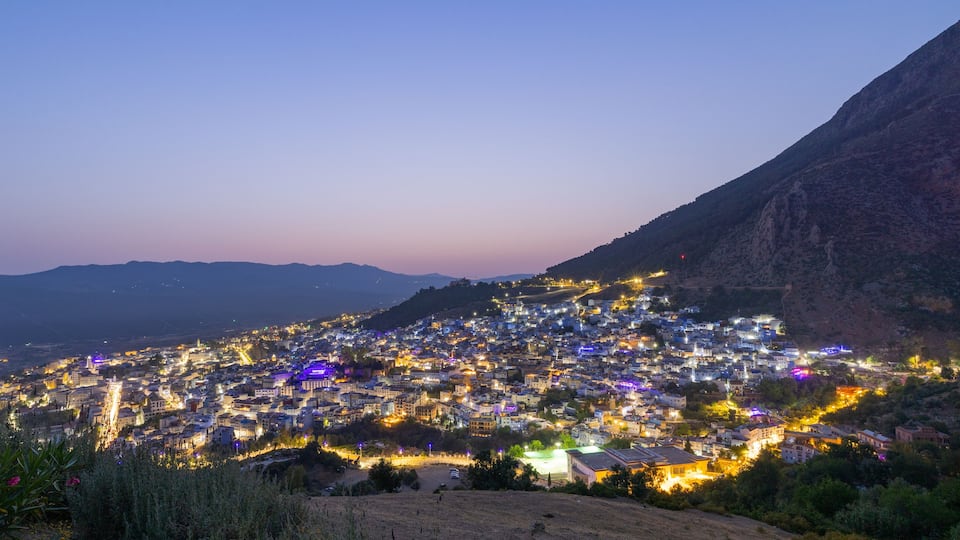 A city at dusk with illuminated buildings and a mountainous backdrop.