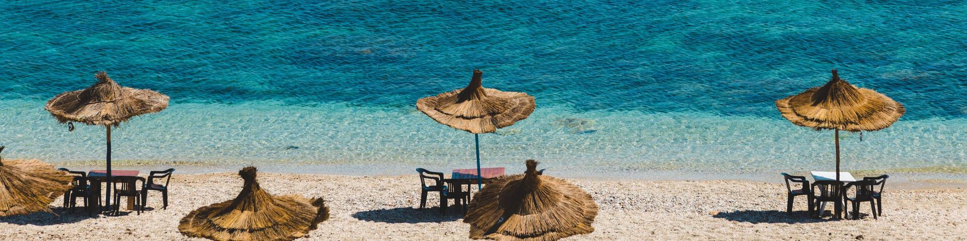 A beach with clear turquoise water, straw umbrellas, and tables set up for dining.