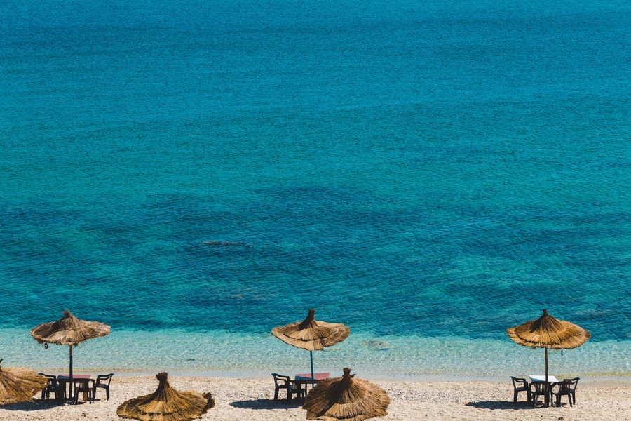 Una playa con agua turquesa cristalina, sombrillas de paja y mesas dispuestas para comer.
