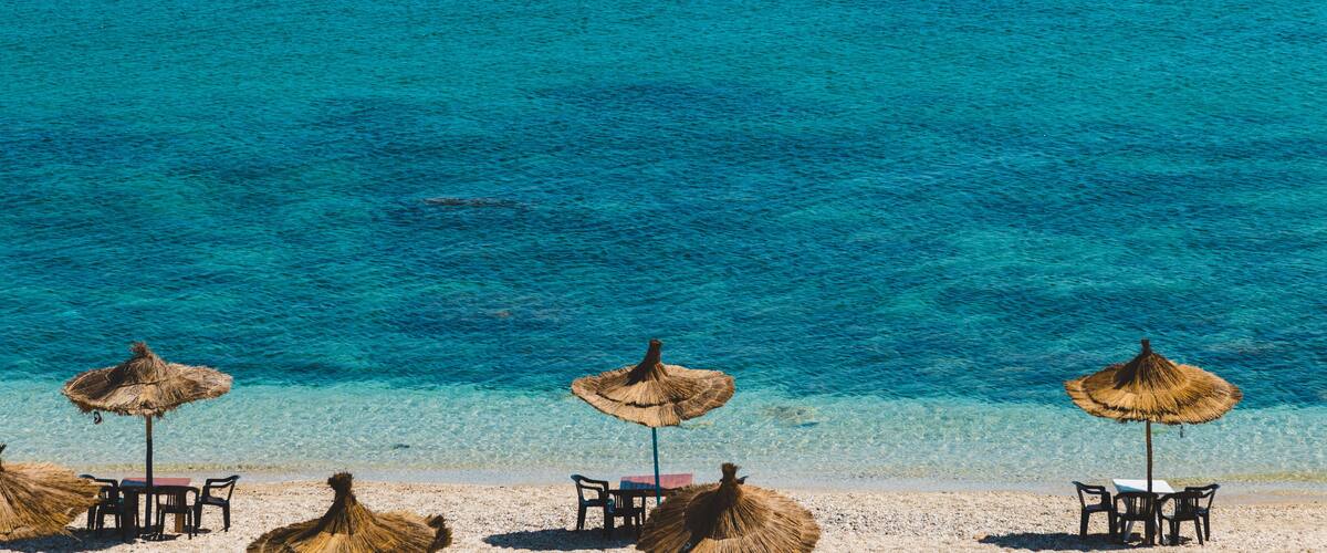 A beach with clear turquoise water, straw umbrellas, and tables set up for dining.