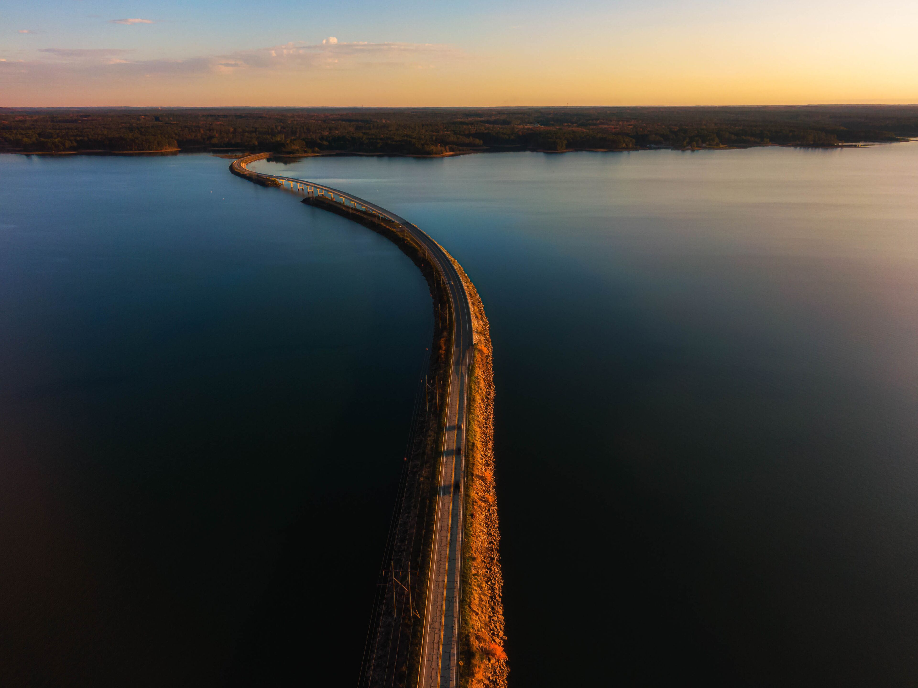 A long bridge curving through a calm body of water, surrounded by forested land.