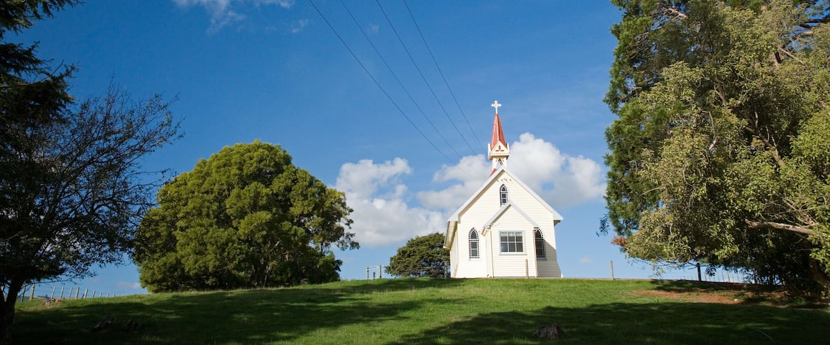 A white church with a red steeple on a grassy hill, surrounded by trees and a clear blue sky.