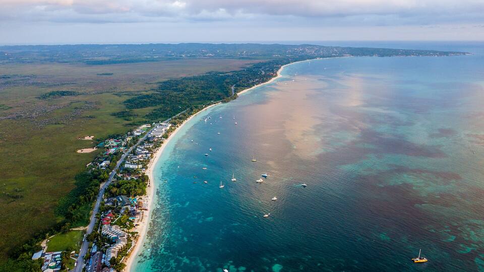 An aerial view of a coastal area with a long sandy beach, clear blue waters, and several boats.