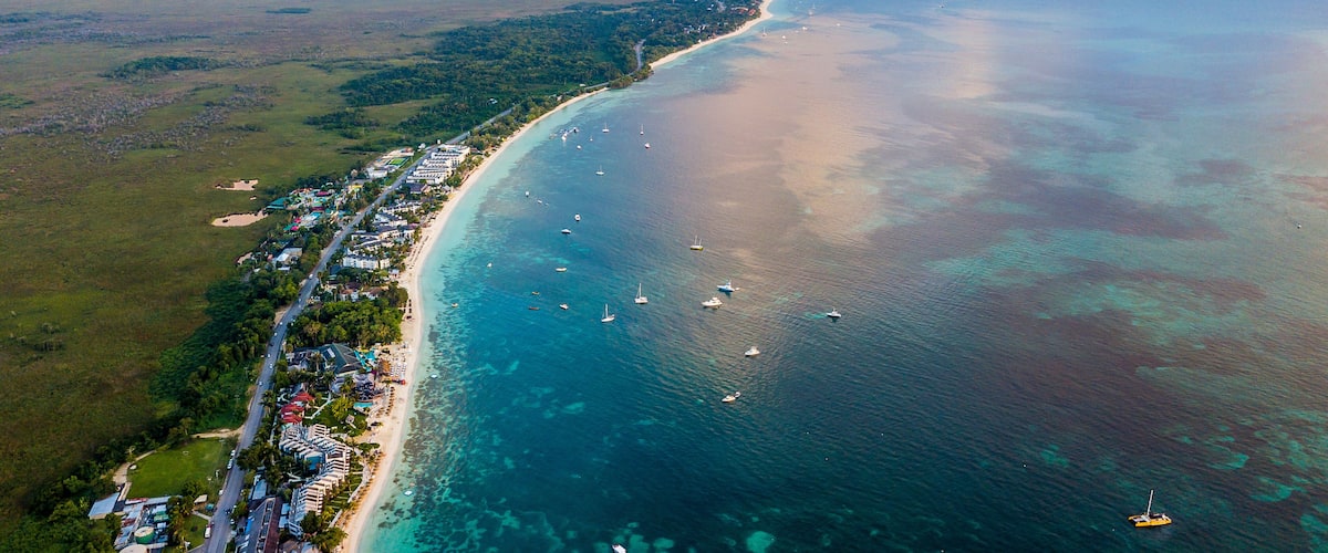 An aerial view of a coastal area with a long sandy beach, clear blue waters, and several boats.