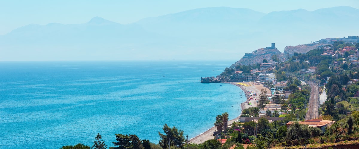 A coastal town with a beach, mountains in the background, and a castle-like structure on a hill.