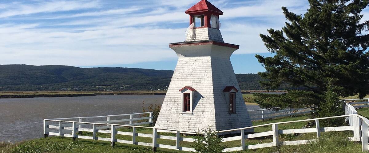 A white lighthouse with a red roof by a lake, surrounded by a white fence and a large tree.
