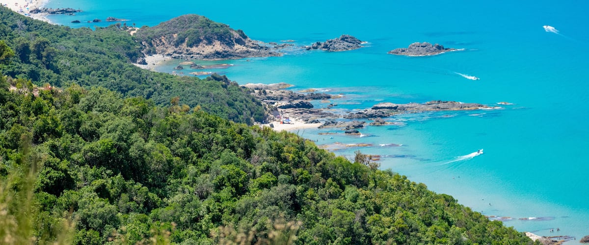 A coastal landscape with a turquoise sea, rocky outcrops, and lush green hills.