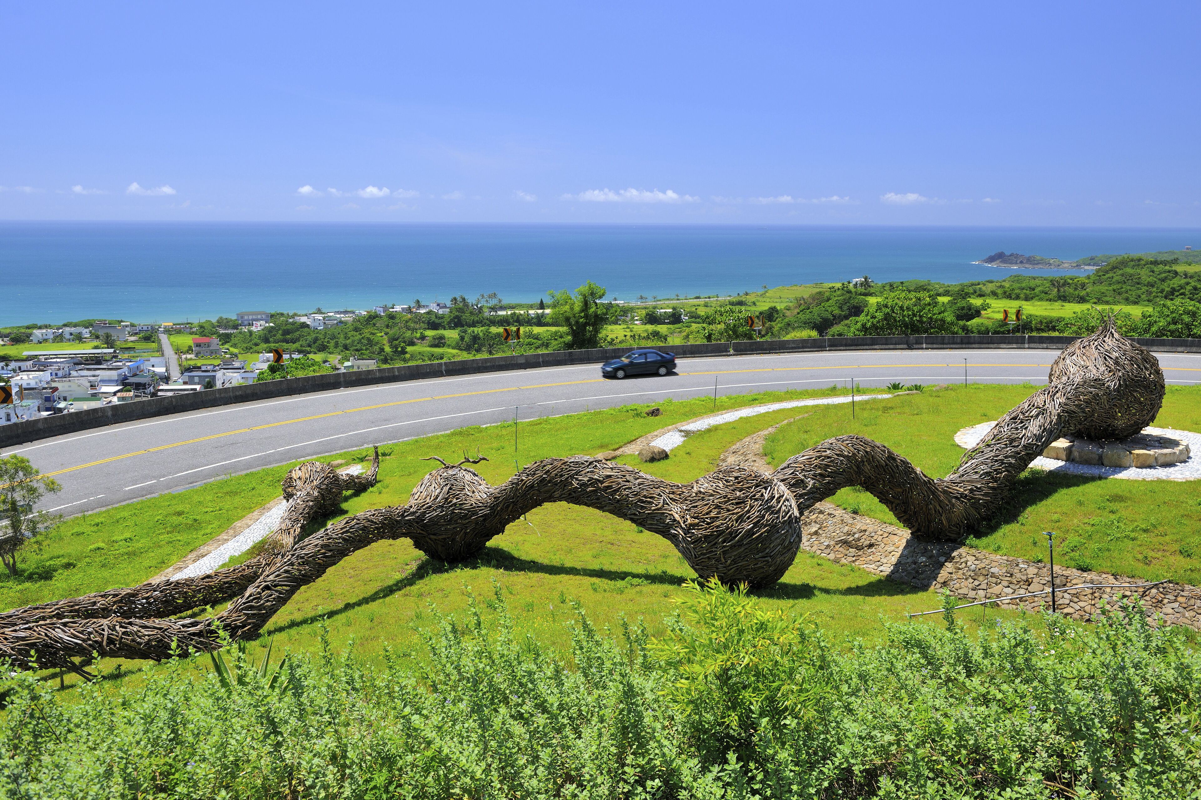 A winding sculpture in a grassy area with a road and ocean in the background.