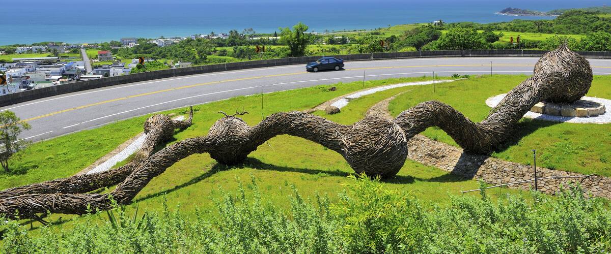 A winding sculpture in a grassy area with a road and ocean in the background.