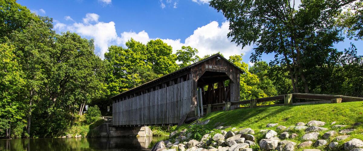 A wooden covered bridge over a calm river, surrounded by lush greenery and rocks.