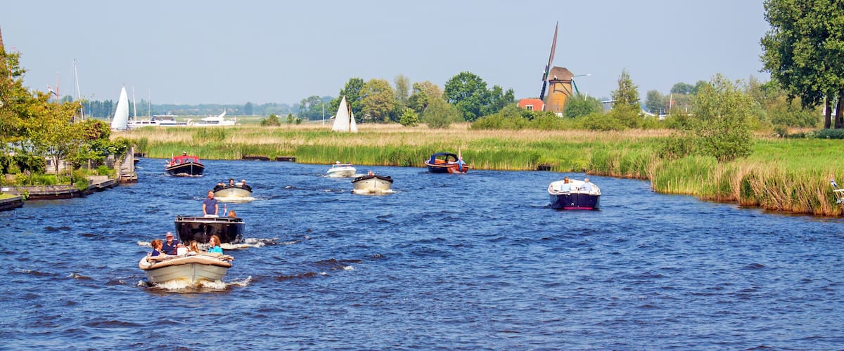 A river with several boats, a windmill, and a sailboat in the background.