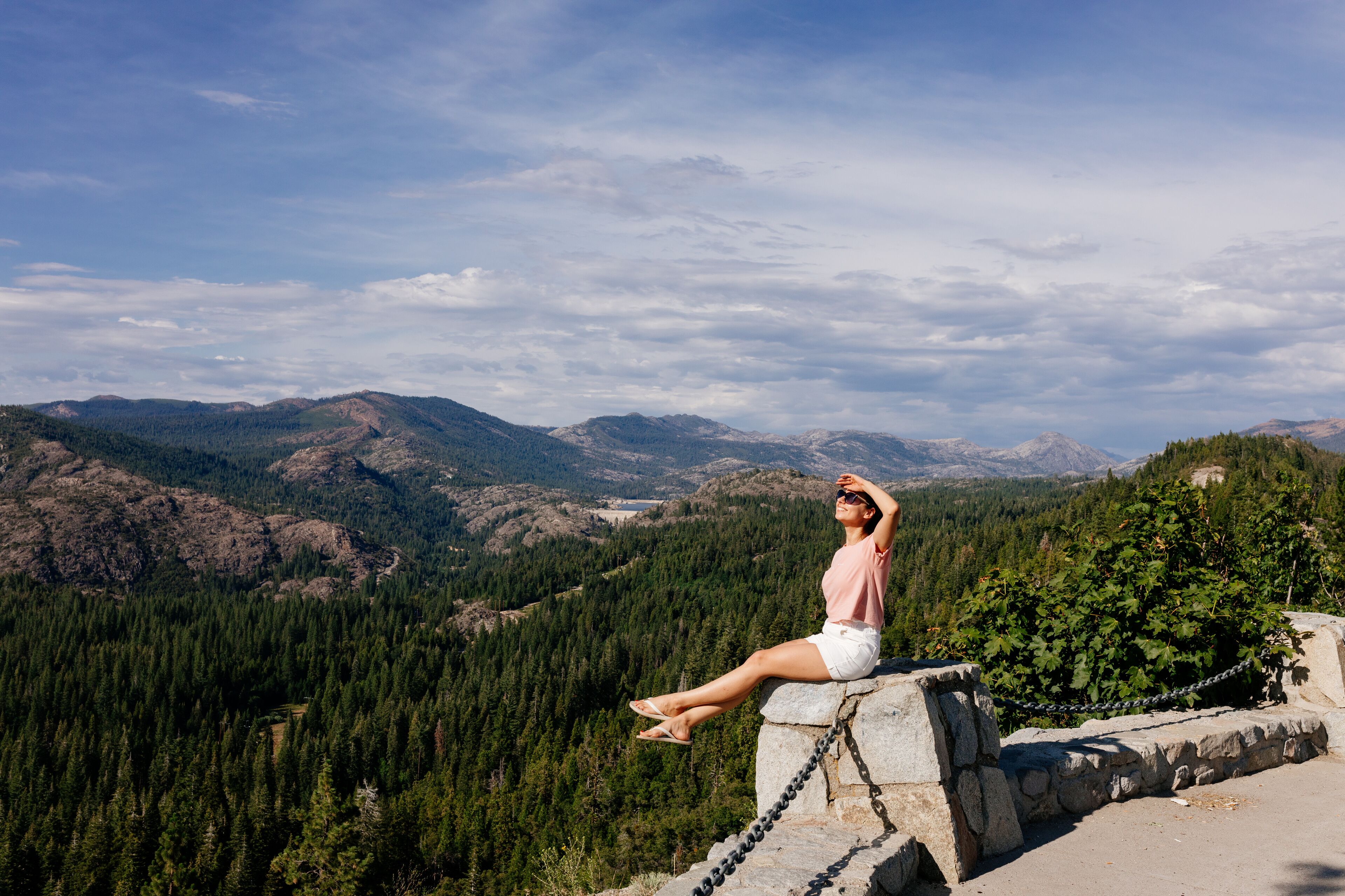 A woman sitting on a stone ledge overlooking a mountainous landscape with dense forests and a clear sky.