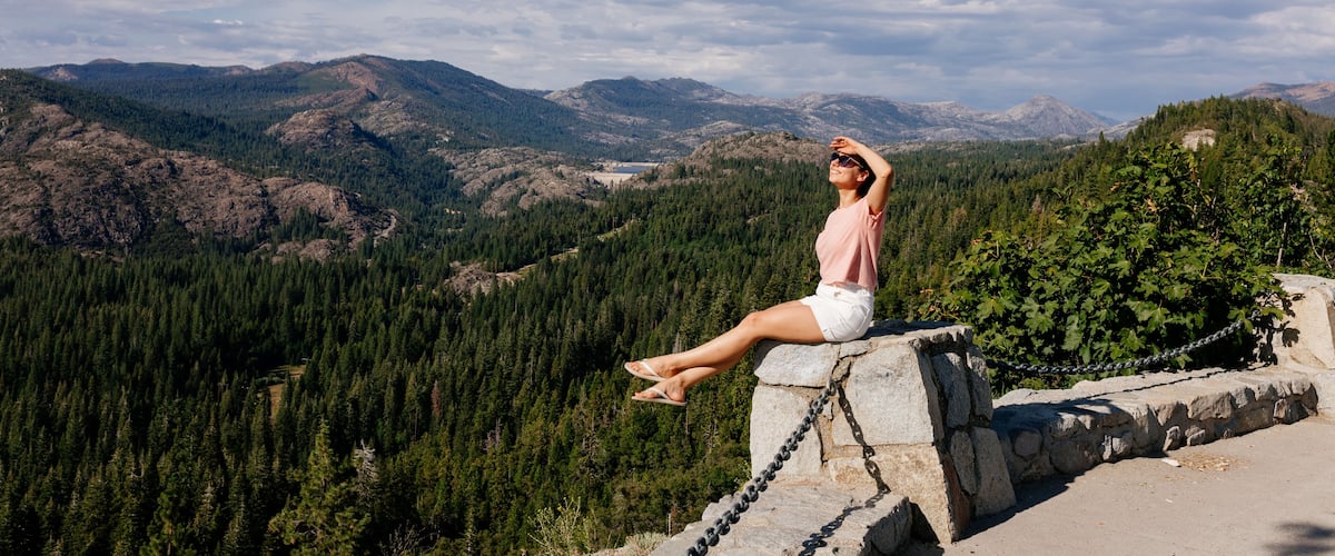 A woman sitting on a stone ledge overlooking a mountainous landscape with dense forests and a clear sky.