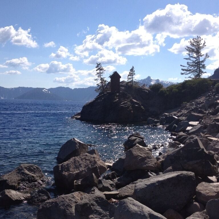 A rocky shoreline with a small wooden structure on a cliff, overlooking a calm sea with mountains in the background.