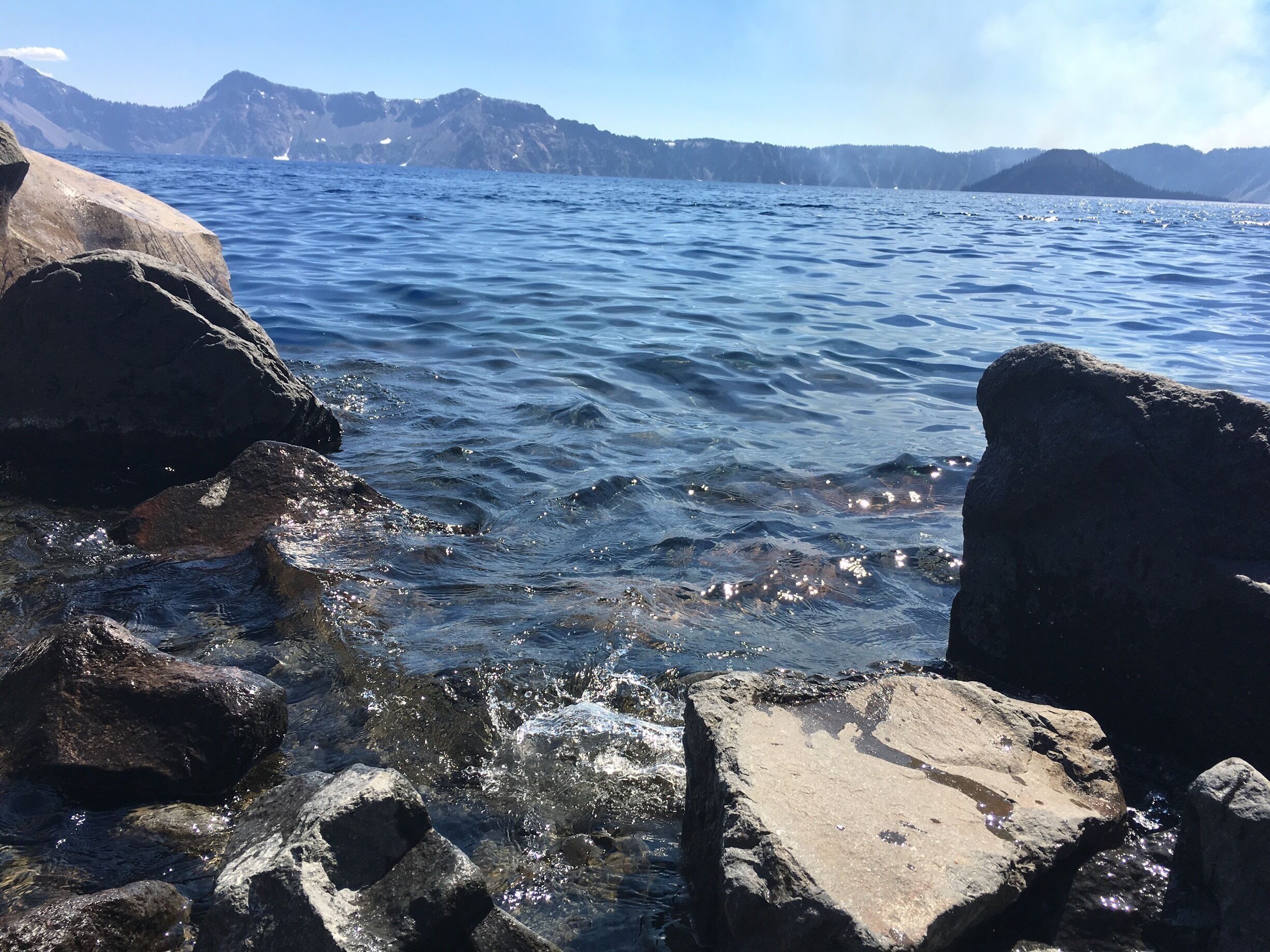 A rocky shoreline with clear blue water and distant mountains.