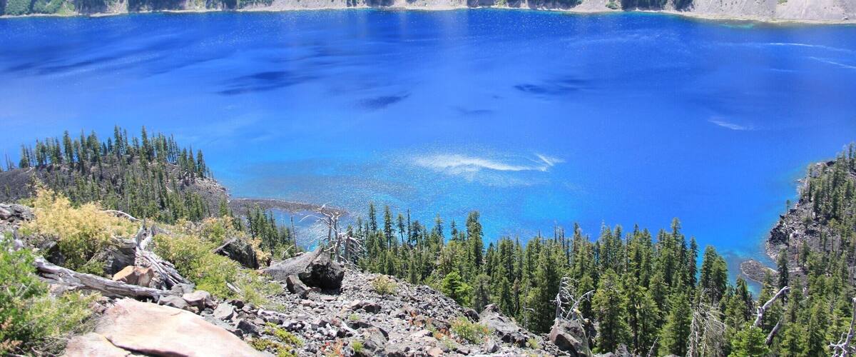 A large, clear blue lake surrounded by dense forests and rocky terrain.