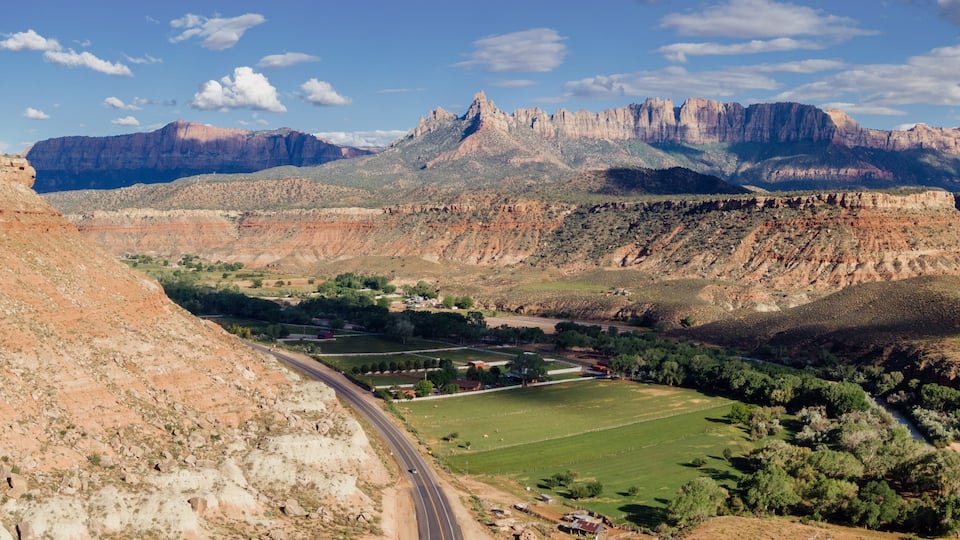 A mountainous landscape with a winding road, green fields, and a prominent rocky peak in the distance.
