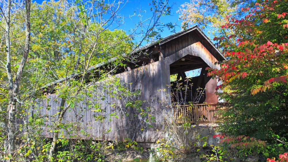 A rustic wooden covered bridge with a steep arch, surrounded by lush greenery and trees.