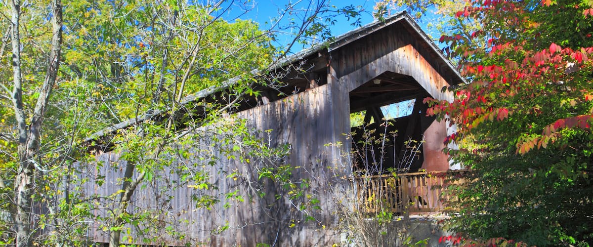A rustic wooden covered bridge with a steep arch, surrounded by lush greenery and trees.