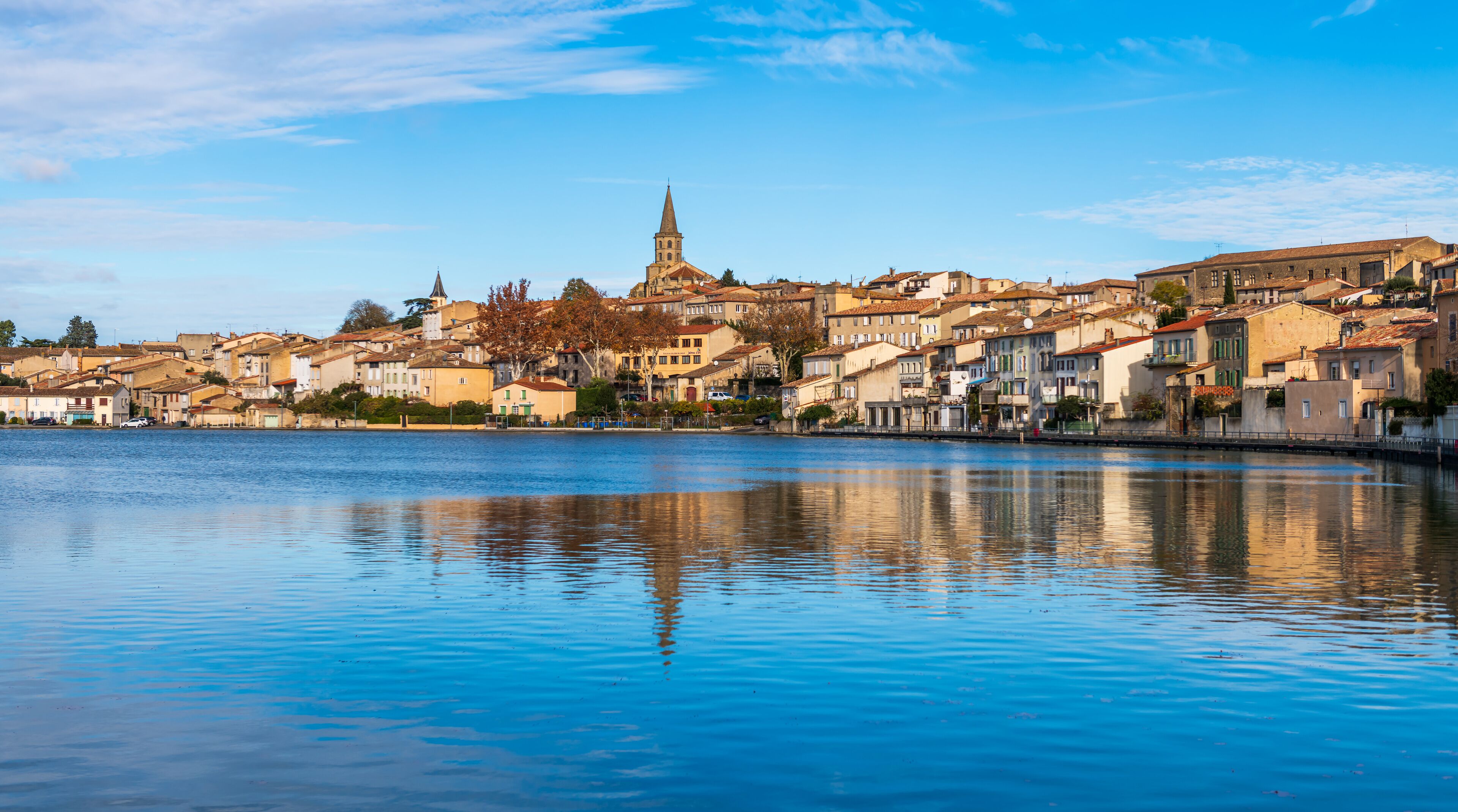 A coastal town with a prominent church tower, calm water reflecting the buildings, and a clear blue sky.