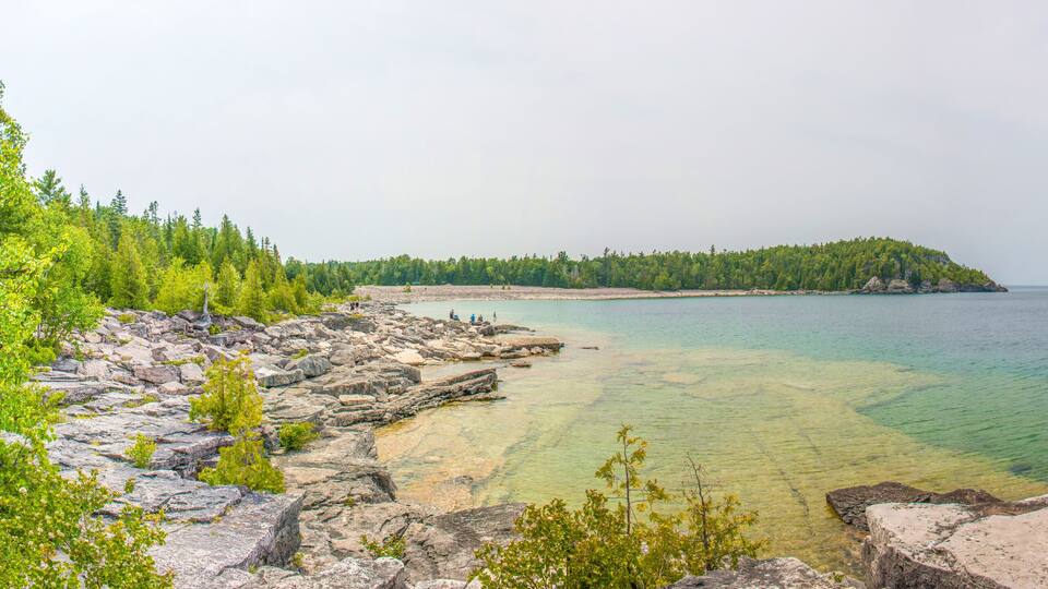 A rocky shoreline with clear water, a forested island, and distant land.
