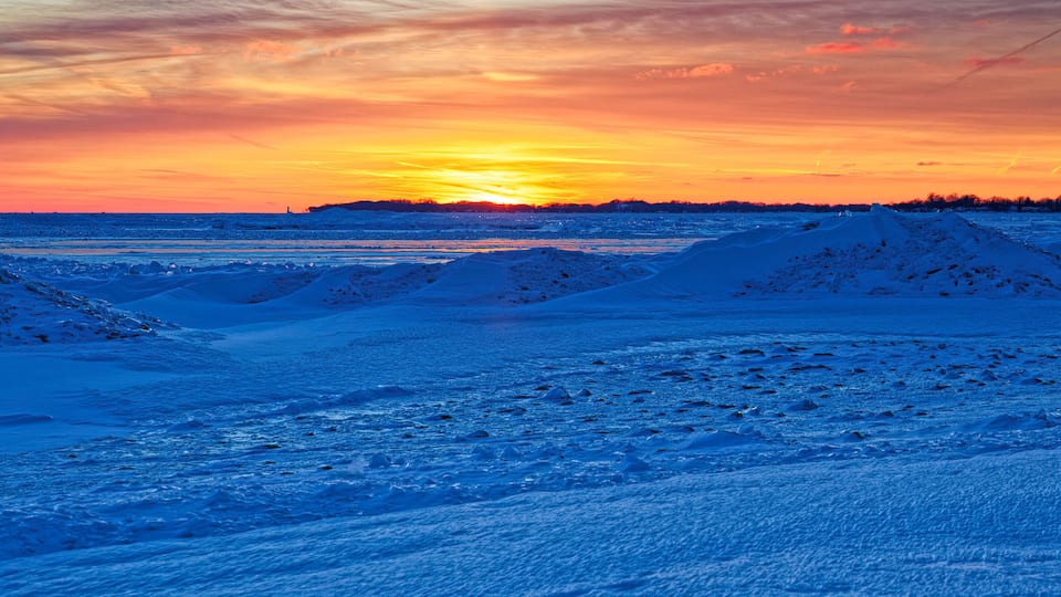 A snowy landscape with a sunset over a frozen body of water, featuring snow-covered dunes and a distant shoreline.