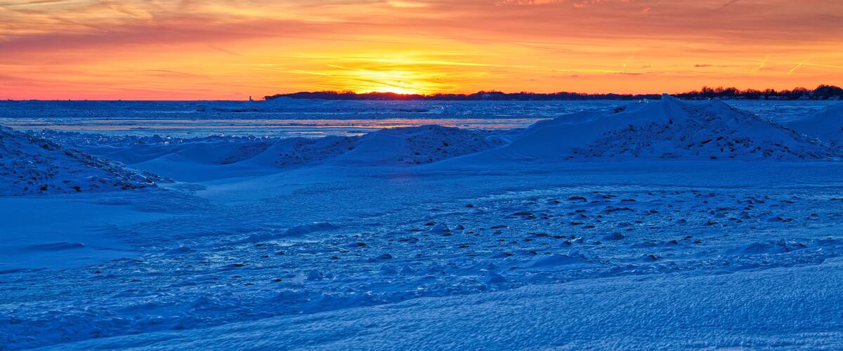 A snowy landscape with a sunset over a frozen body of water, featuring snow-covered dunes and a distant shoreline.