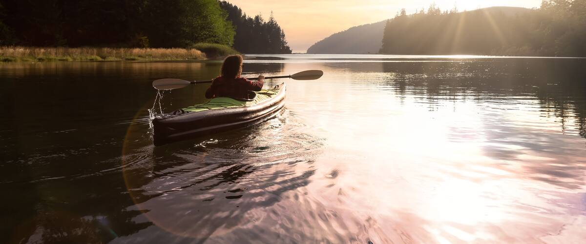 A person kayaking on a calm lake surrounded by dense forest during sunset.