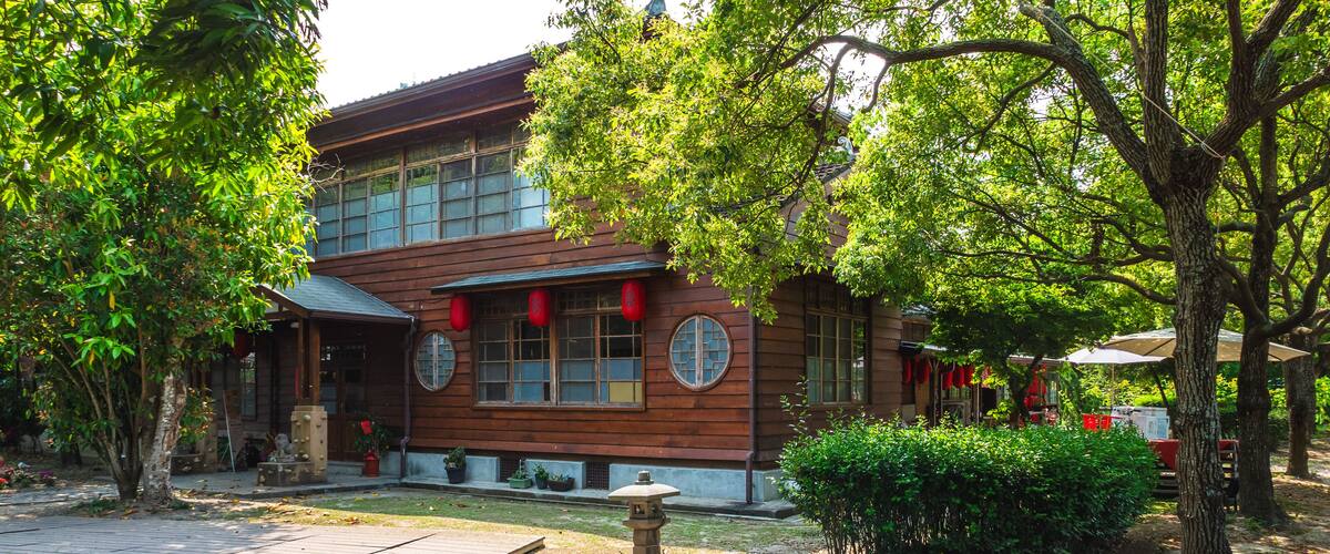 A traditional wooden building with large windows, surrounded by lush greenery and a stone pathway.