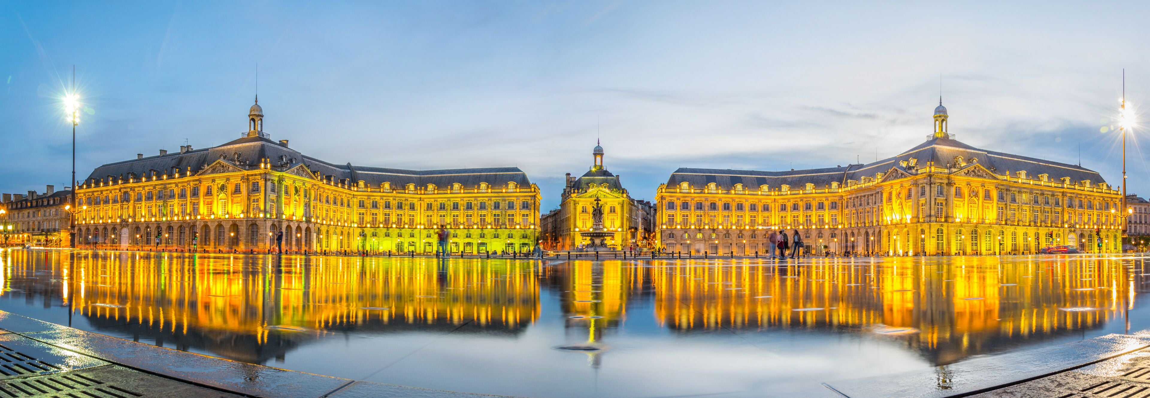 A grand, illuminated building with a central dome and two symmetrical wings, reflecting on a calm water surface.