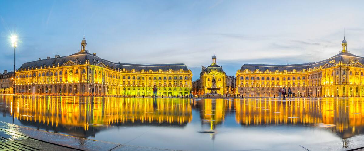A grand, illuminated building with a central dome and two symmetrical wings, reflecting on a calm water surface.