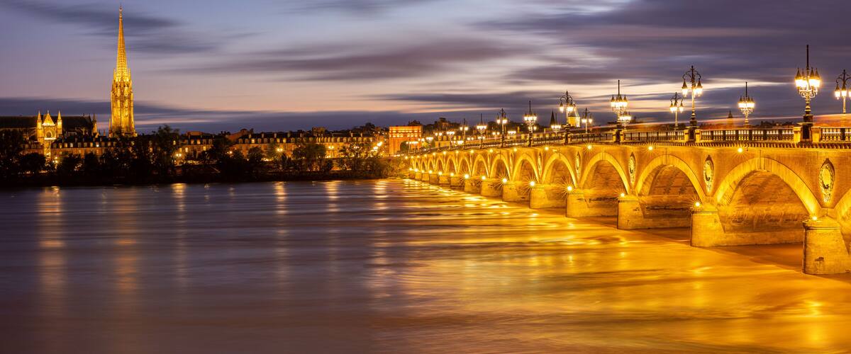 A lit bridge with arches over a river, a tall cathedral spire, and city buildings in the background.
