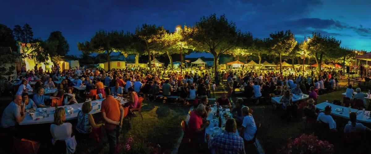 A crowded outdoor event with tables and people under trees at dusk.