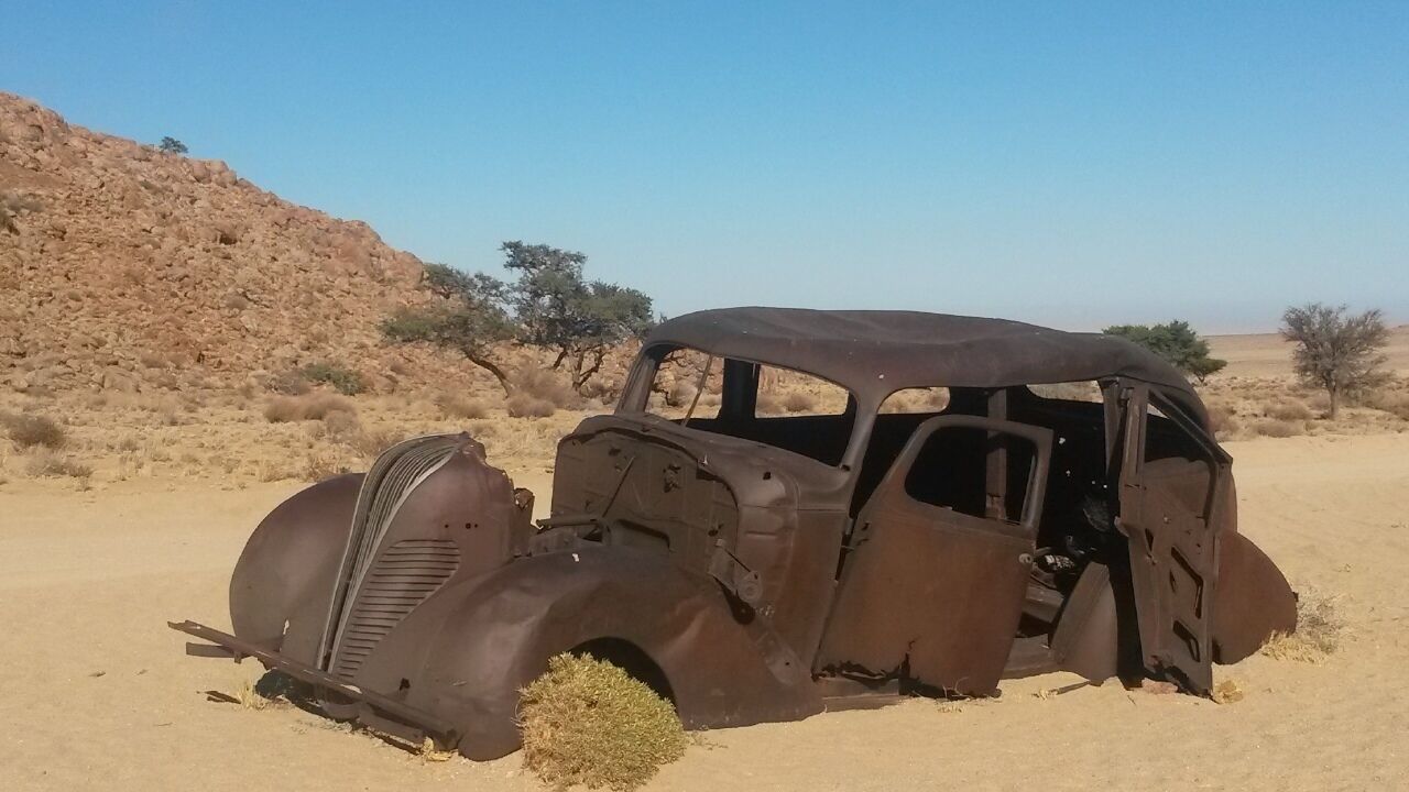 An old, rusted car in a desert with sparse vegetation and rocky hills.