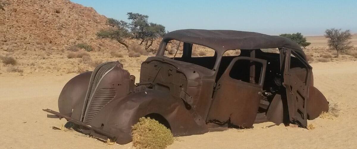 An old, rusted car in a desert with sparse vegetation and rocky hills.