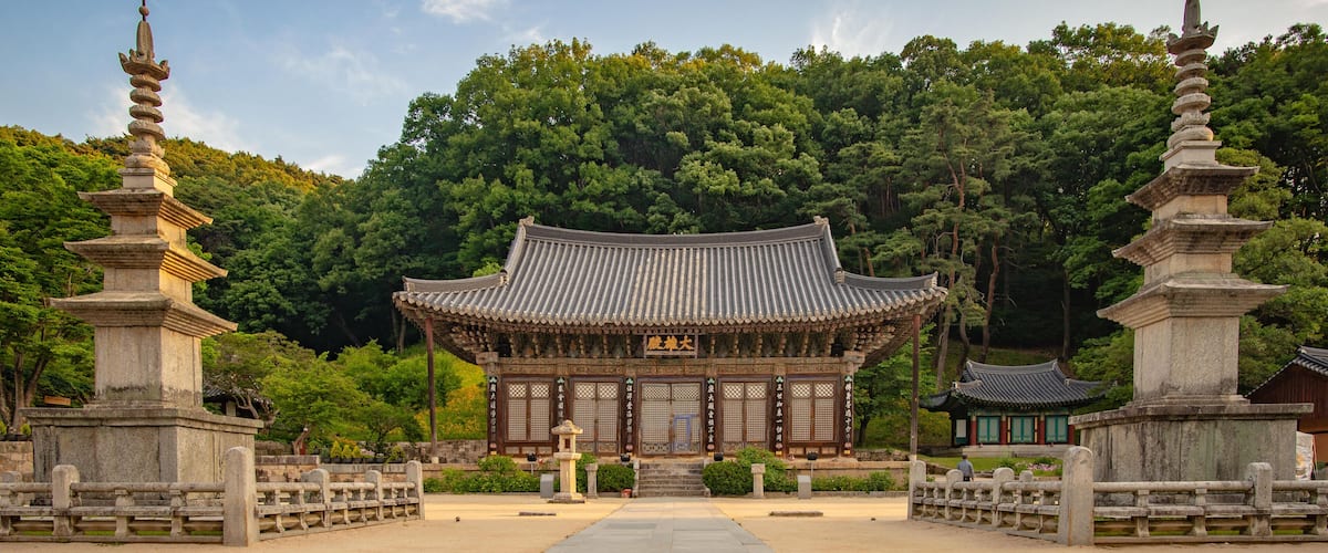 A traditional Japanese temple with a central building, surrounded by stone lanterns and a forested hillside.