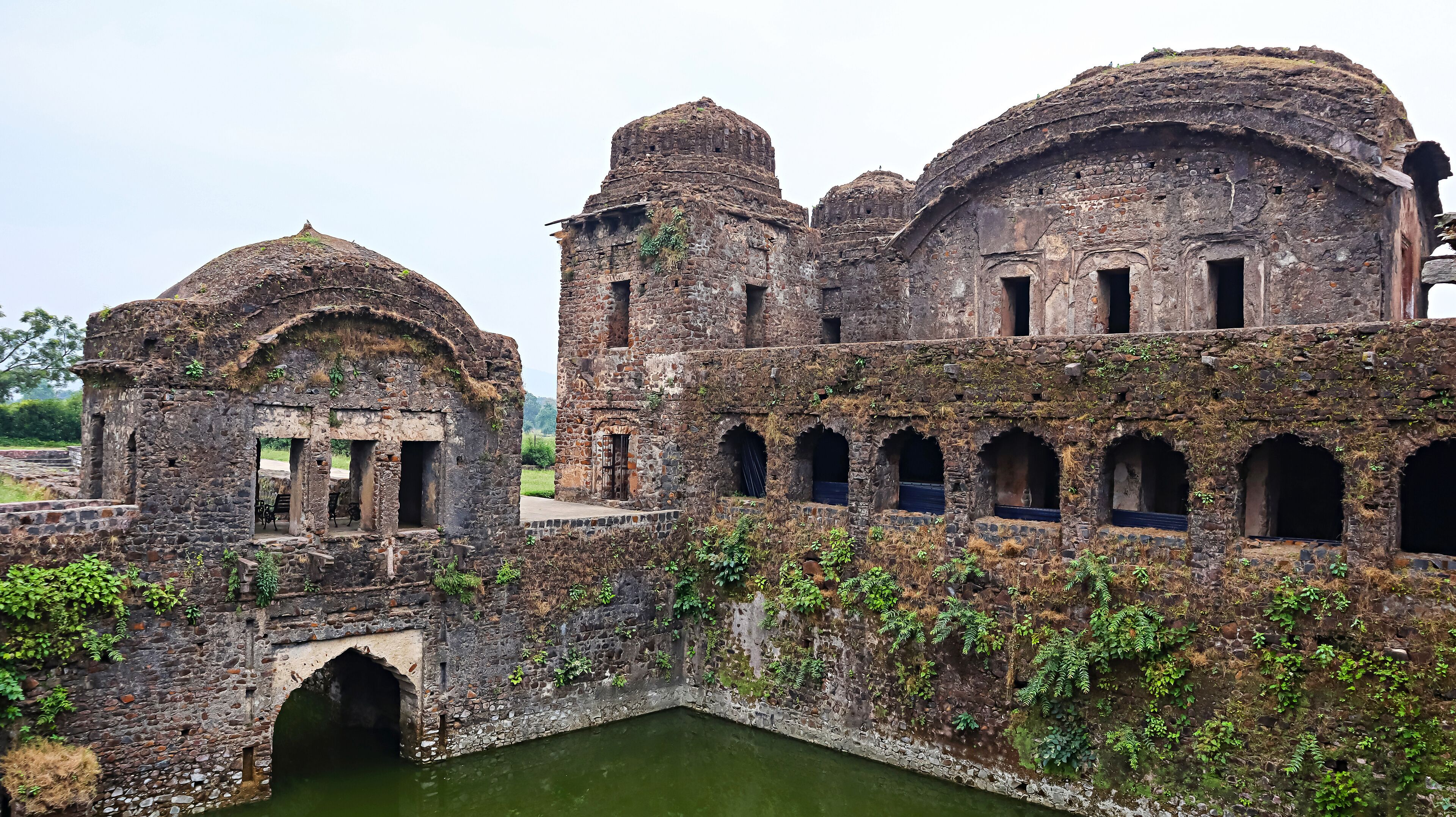 An ancient stone structure with domed roofs and arched windows, partially overgrown with vegetation.