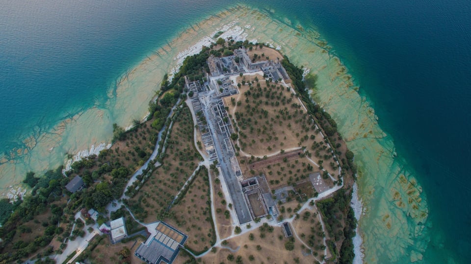 An aerial view of a coastal area with a clear beach, a large building complex, and surrounding greenery.