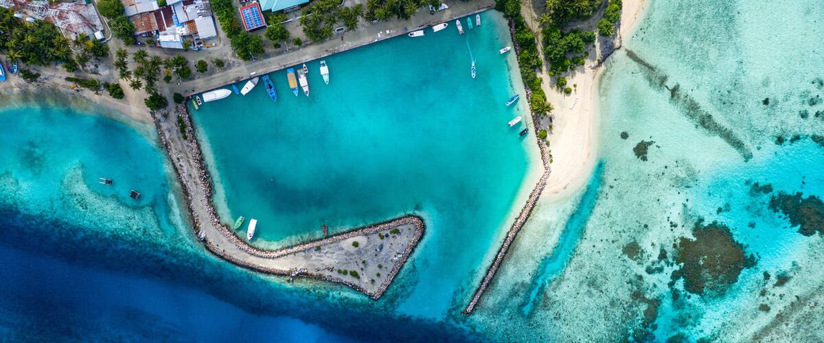 An aerial view of a coastal area with a clear blue lagoon, a small island with a rocky perimeter, and a beach with boats and a pier.