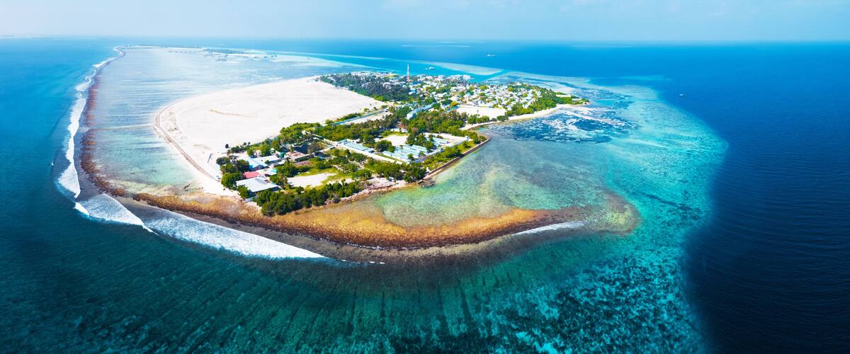 An aerial view of a coastal island with a sandy beach, clear turquoise waters, and a small settlement with buildings and greenery.