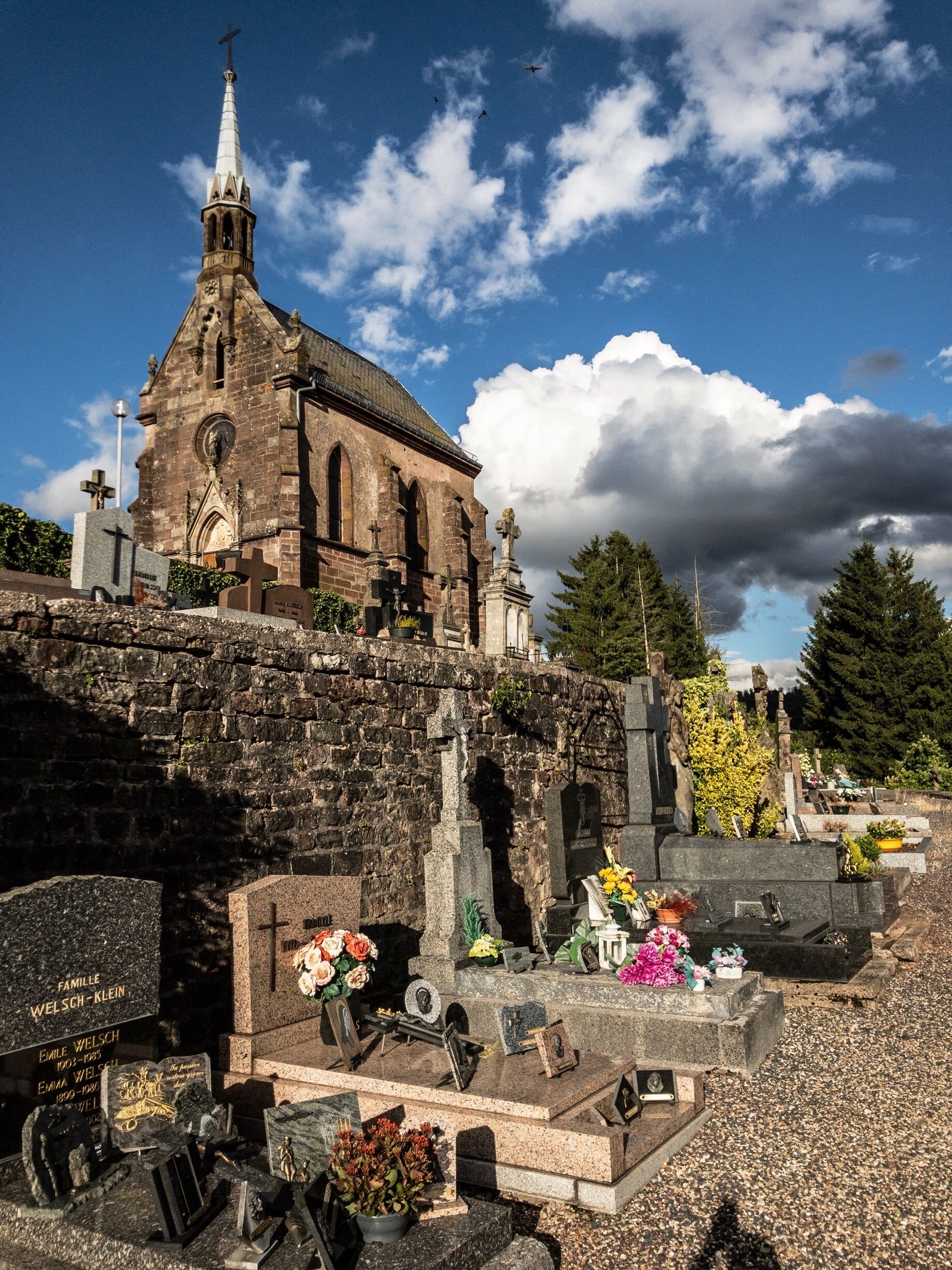 A church with a tall steeple and a cemetery with gravestones and flowers.