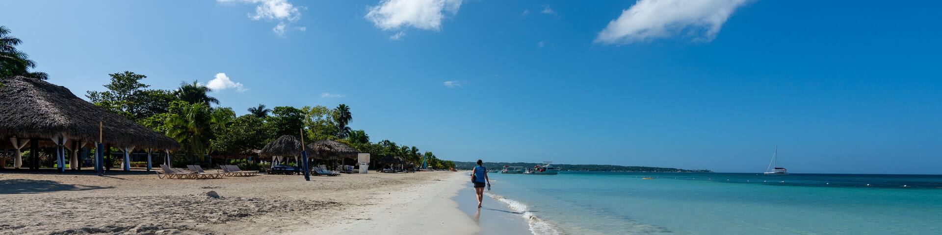 A beach with clear turquoise water, a person walking along the shore, and thatched-roof structures on the left.