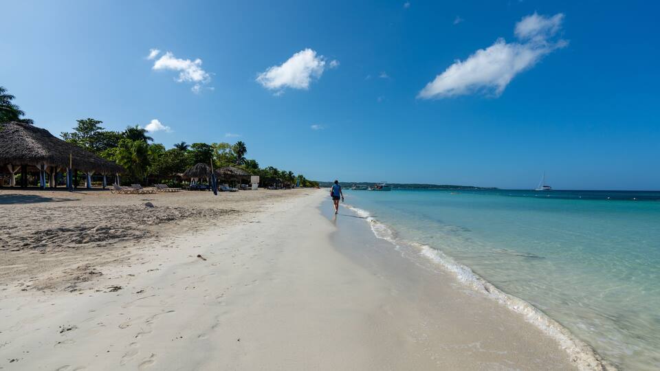 A beach with clear turquoise water, a person walking along the shore, and thatched-roof structures on the left.
