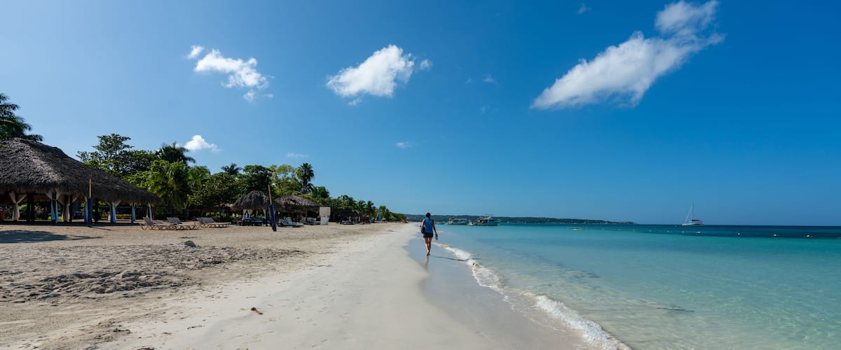 A beach with clear turquoise water, a person walking along the shore, and thatched-roof structures on the left.