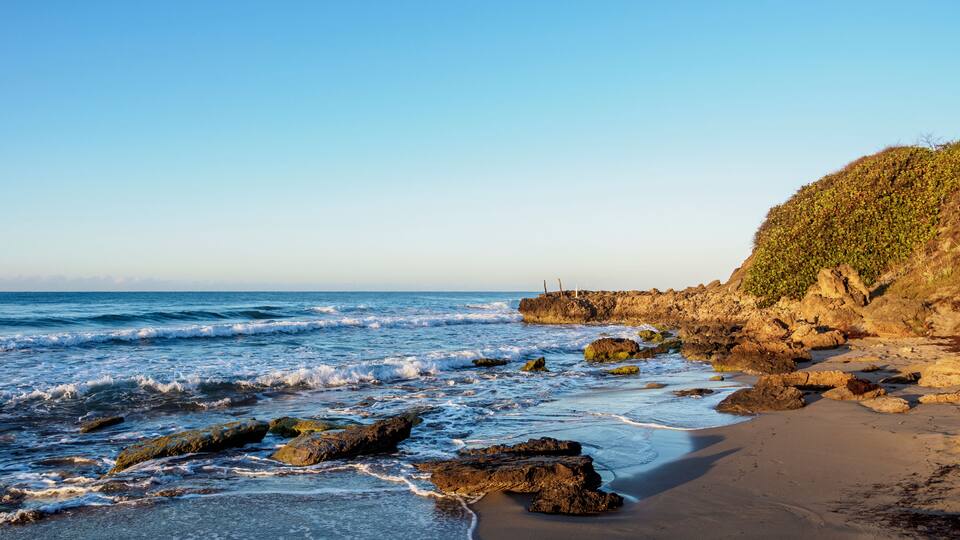 A rocky coastline with waves, a sandy beach, and a hill with vegetation.