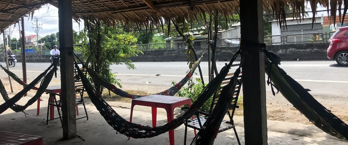 A shaded outdoor seating area with hammocks and red plastic chairs, overlooking a road with passing vehicles.